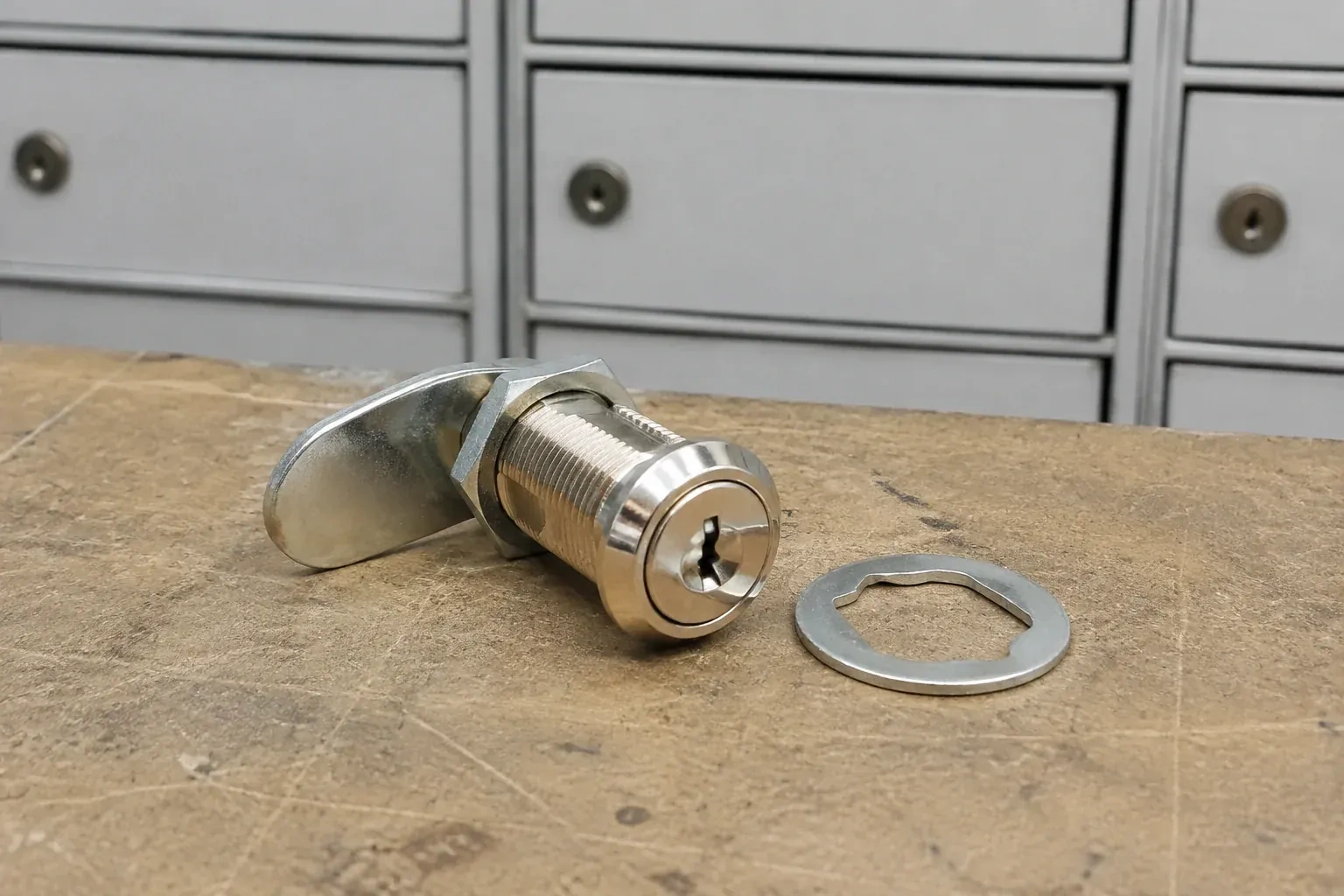 Detached threaded cam lock and mounting washer on a workbench in front of closed mail compartments. — Mailbox Lock Replacement, Lincoln