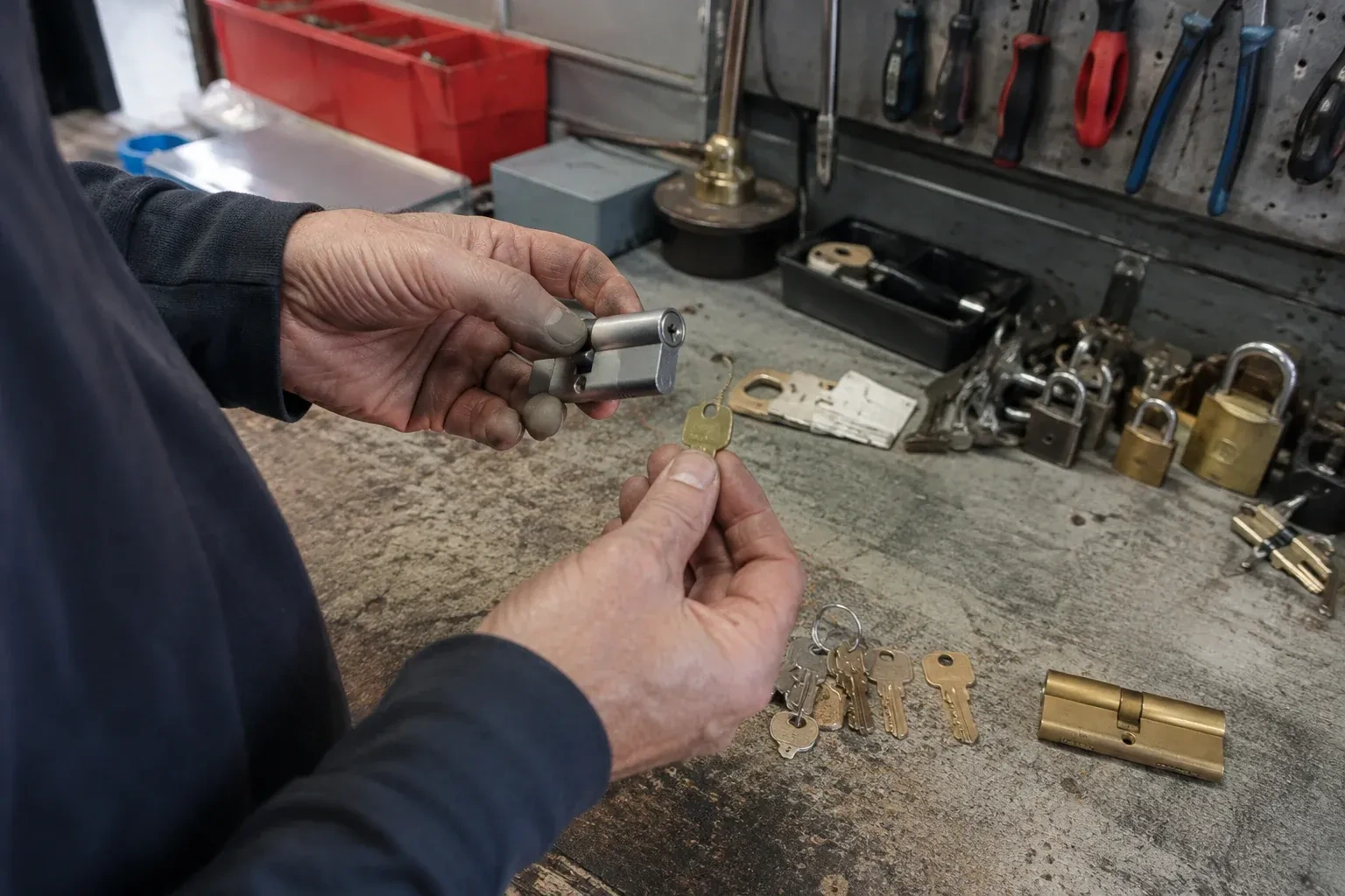 Hands holding a silver-colored lock cylinder and a brass-colored key above a workbench with padlocks, keys and tools. — Master Key System Design, Lincoln